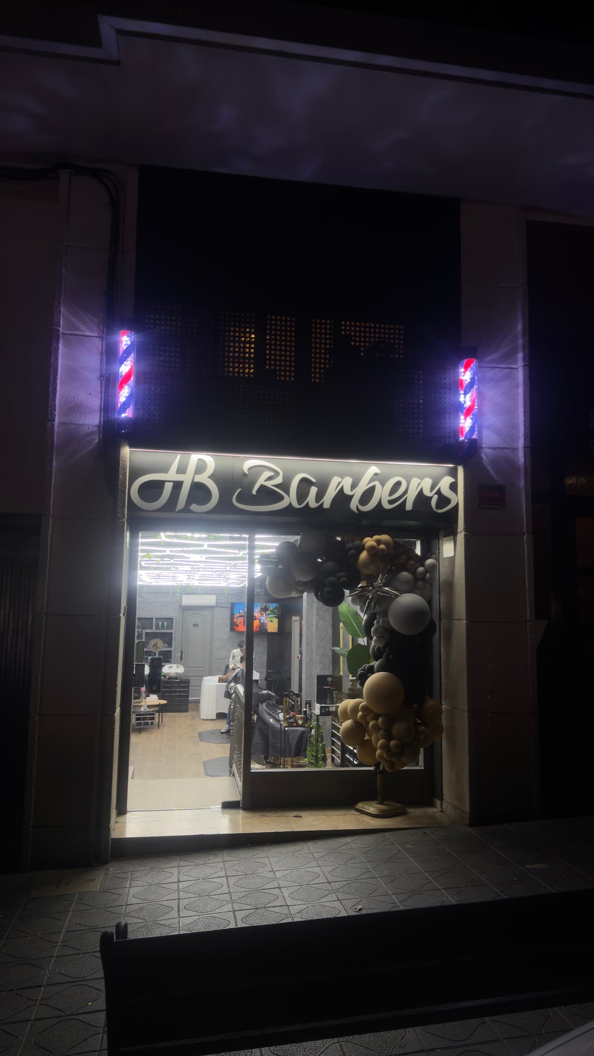 Fachada nocturna de HB Barbers con el rótulo iluminado y el poste de barbero clásico en la entrada